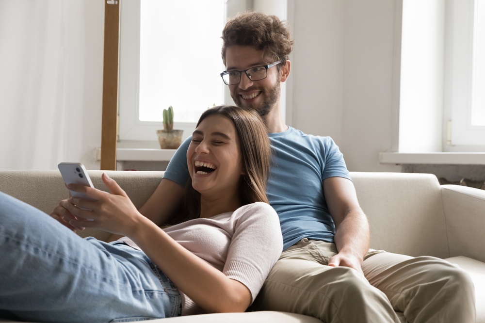 A happy couple laughing together on the couch, representing comfortable, confident smiles achieved through quality dental care – Dentist in Redondo Beach.