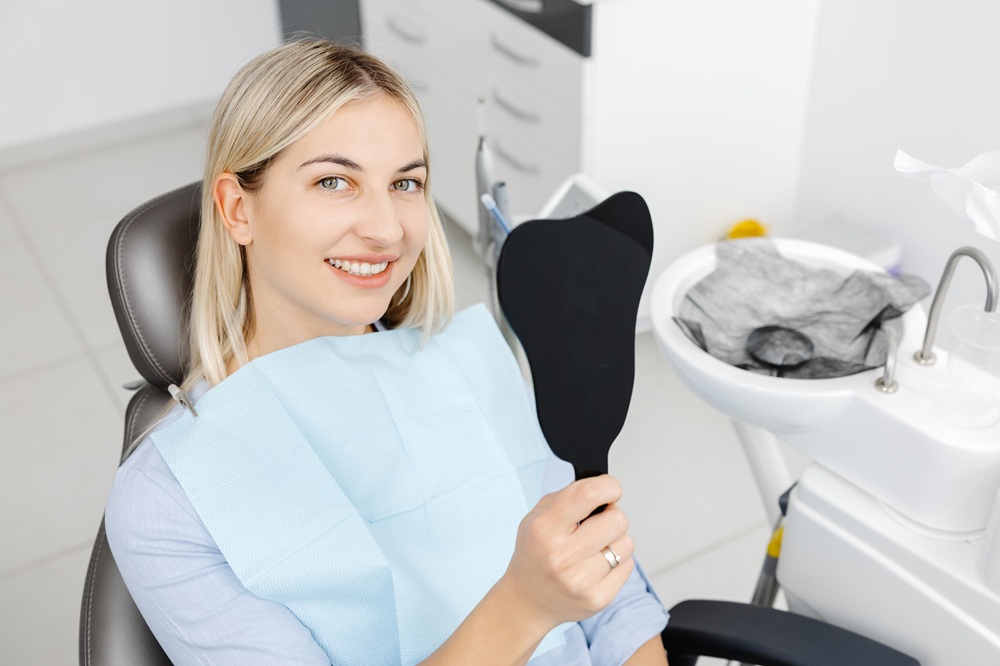 A happy patient sits in a modern dental chair holding a mirror and admiring her bright, freshly cleaned smile after a professional dental visit – Redondo Beach Dental Cleanings