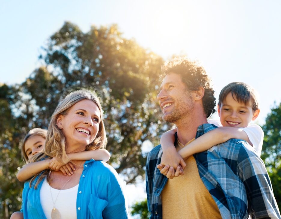 Smiling family enjoying outdoor time together – Palos Verdes Dental Care A happy family of four enjoys a sunny day outdoors, with two children playfully riding on their parents’ backs, symbolizing joy, togetherness, and the importance of a healthy smile – Palos Verdes Dental Care